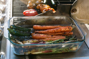 Glass dish filled with grilled carrots, scallions, and other charred vegetables, sitting on a metal outdoor table.