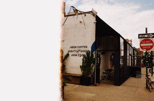 Street corner café with “Fresh Coffee, Quality Goods, Local Food” painted on the wall, black awning and patio setup beside “Do Not Enter” sign.