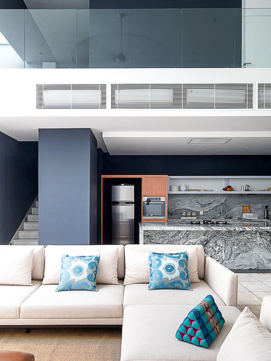 Ruang tamu modern dengan sofa krem berpadukan bantal biru, dapur marmer, tangga, dan balkon kaca di latar.