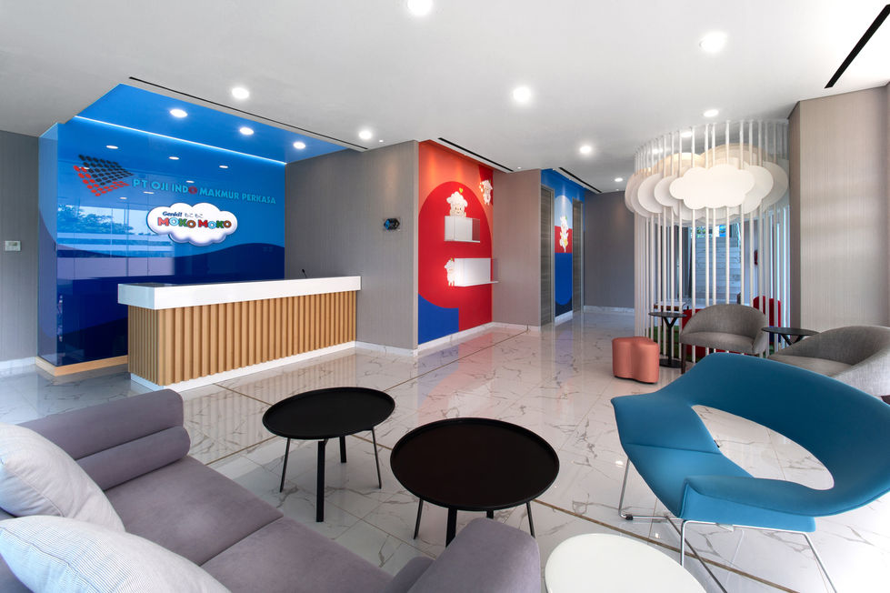 Modern corporate lobby with a blue Moko Moko branded reception wall, slatted wood desk, grey sofas, a blue sculptural armchair, and decorative cloud installations.