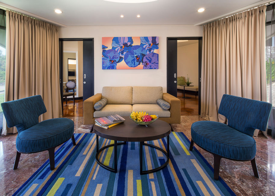 Bright resort suite living room featuring a tan sofa, blue accent chairs, a colorful striped rug, and floral wall art.