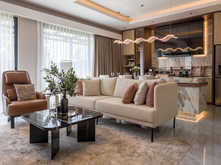 A modern open-concept living and dining area with warm, ambient lighting. In the foreground, a cream-colored sofa with accent pillows sits on a neutral area rug facing a dark marble coffee table. To the left sits a tan leather armchair next to floor-to-ceiling windows with sheer white and beige drapes. In the background, a luxurious kitchen features marble countertops, dark cabinetry, and a wavy LED pendant light hanging over the kitchen island.