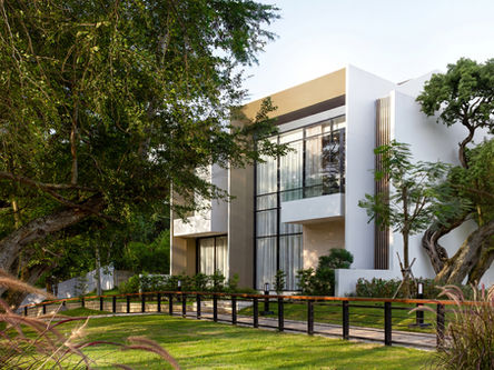 Exterior view of a contemporary two-story white villa with large floor-to-ceiling glass panel windows, nestled within a lush natural landscape. Broad-canopied trees frame the building, with a gnarled trunk extending near the right facade. In the foreground, a paved pathway flanked by a low wooden railing runs alongside a green lawn and ornamental fountain grasses under bright, natural daylight.