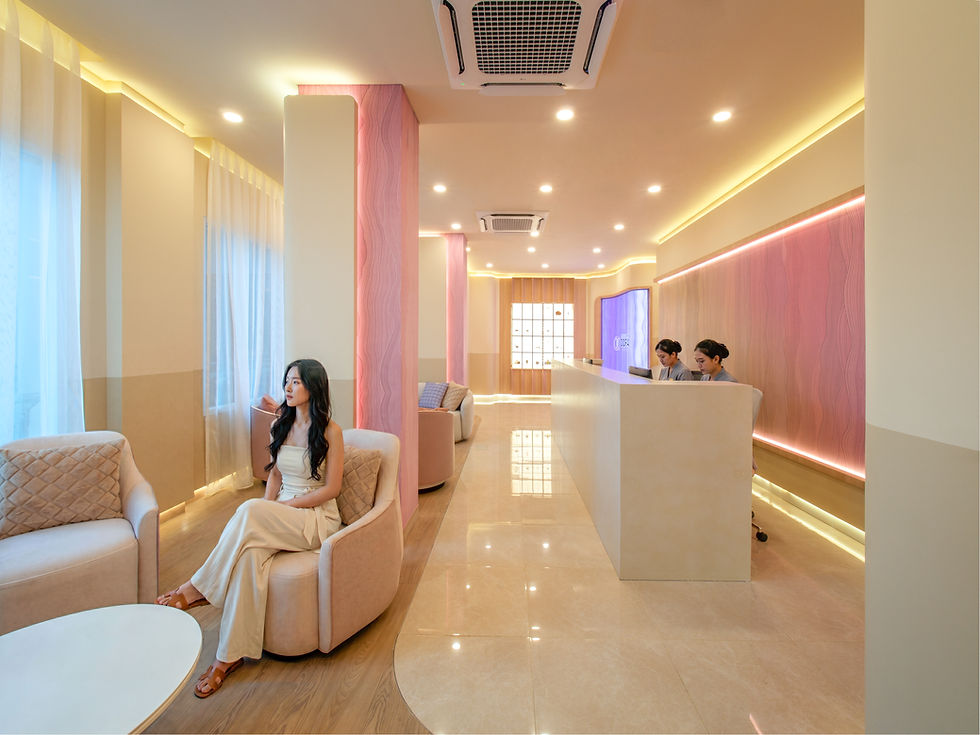 Woman in cream dress sits in a Oora Skin Labs Lobby Reception Lounge area while two staff work behind a reception desk, with soft pink lighting.
