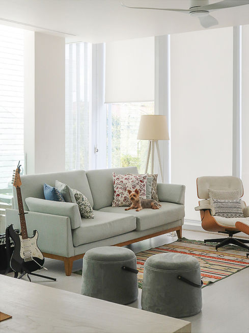 Cozy living room area featuring a light blue sofa with decorative pillows, an armchair, two ottomans, an electric guitar, and bright window blinds.