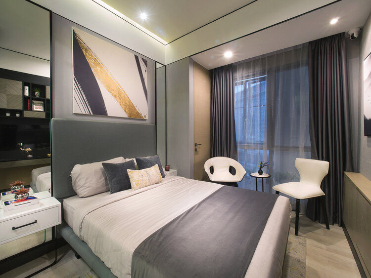 Studio apartment bedroom featuring a grey headboard bed, decorative art panels, dark curtains, and two modern white chairs by the window.