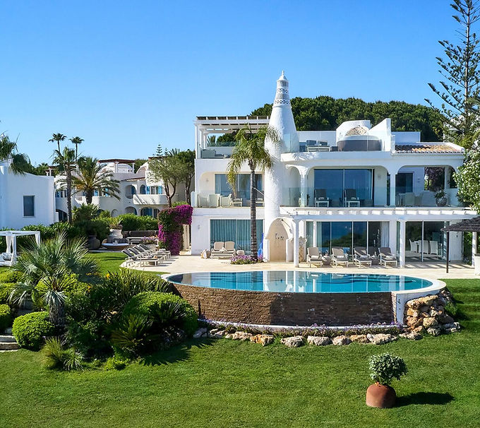 Garden view looking back at a luxury modern while villa with several floors and a round outdoor pool surrounded by green garden