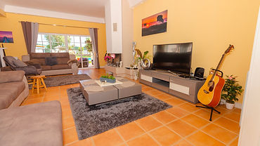 A cozy living room with yellow walls, terracotta tiled floors, a gray sectional sofa, a guitar leaning against a TV stand, and a gray fluffy rug in the center.