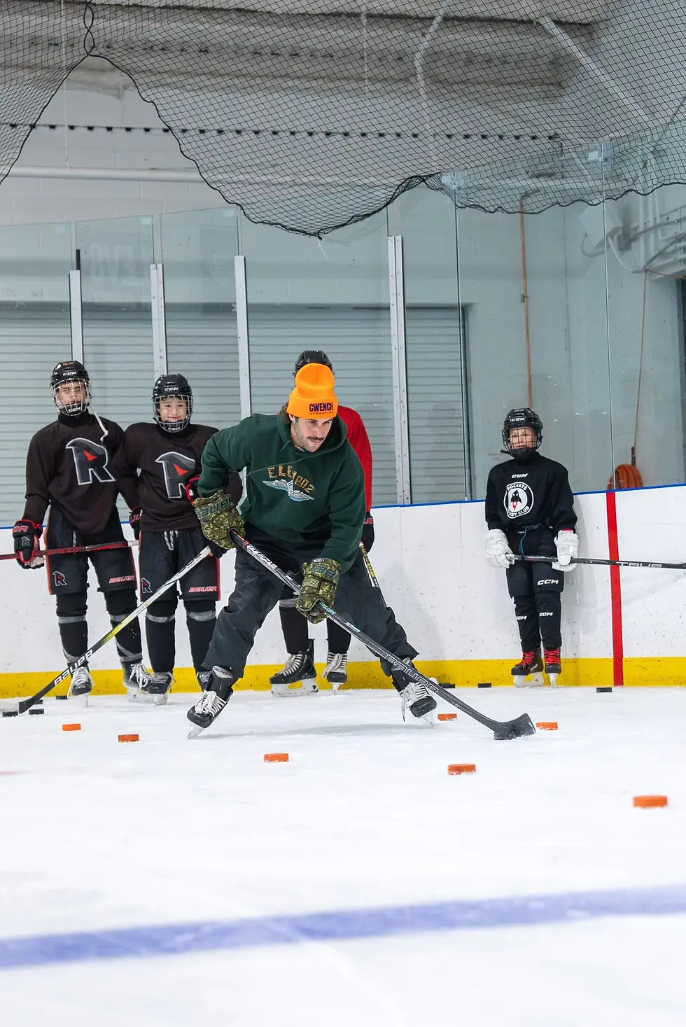 Hockey player in orange hat practicing on ice rink with pucks, while three children watch. Others wear dark uniforms. Indoor setting.