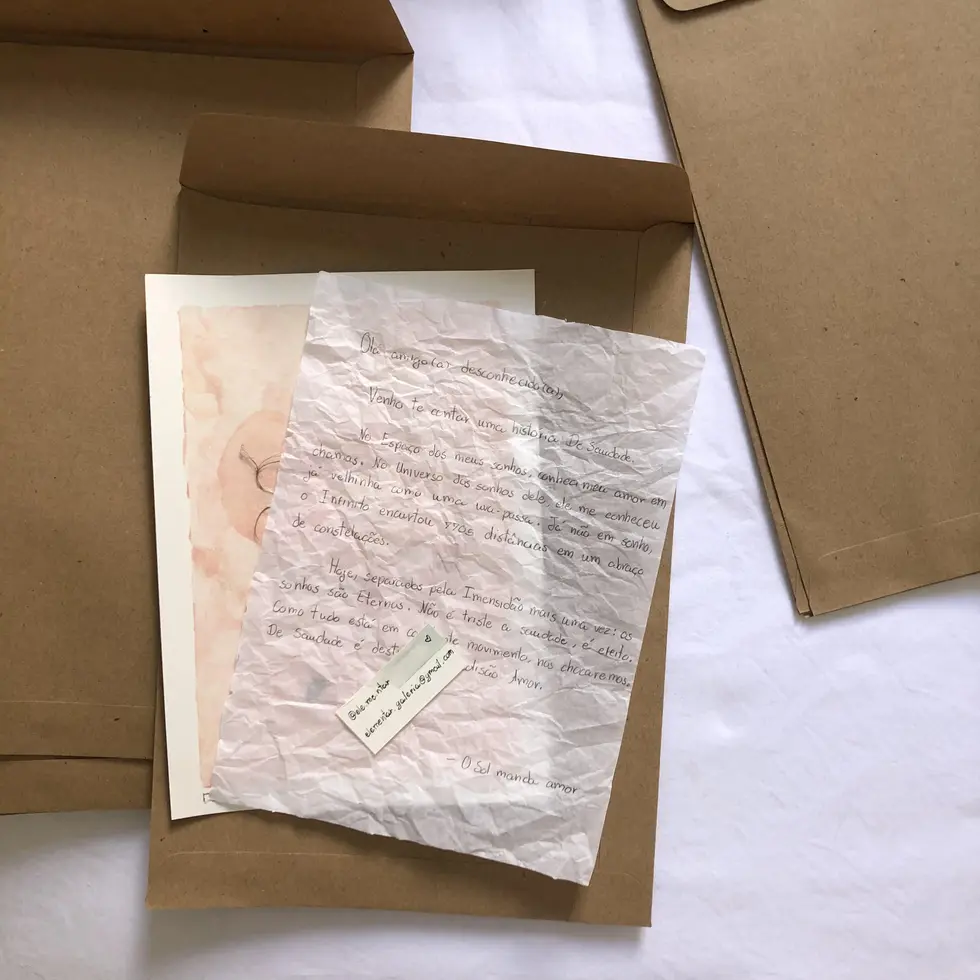 Envelope pardo com folha de papel de seda branca amassada, trazendo carta manuscrita a um destinatário desconhecido e pequeno cartão artesanal com formas de contato.