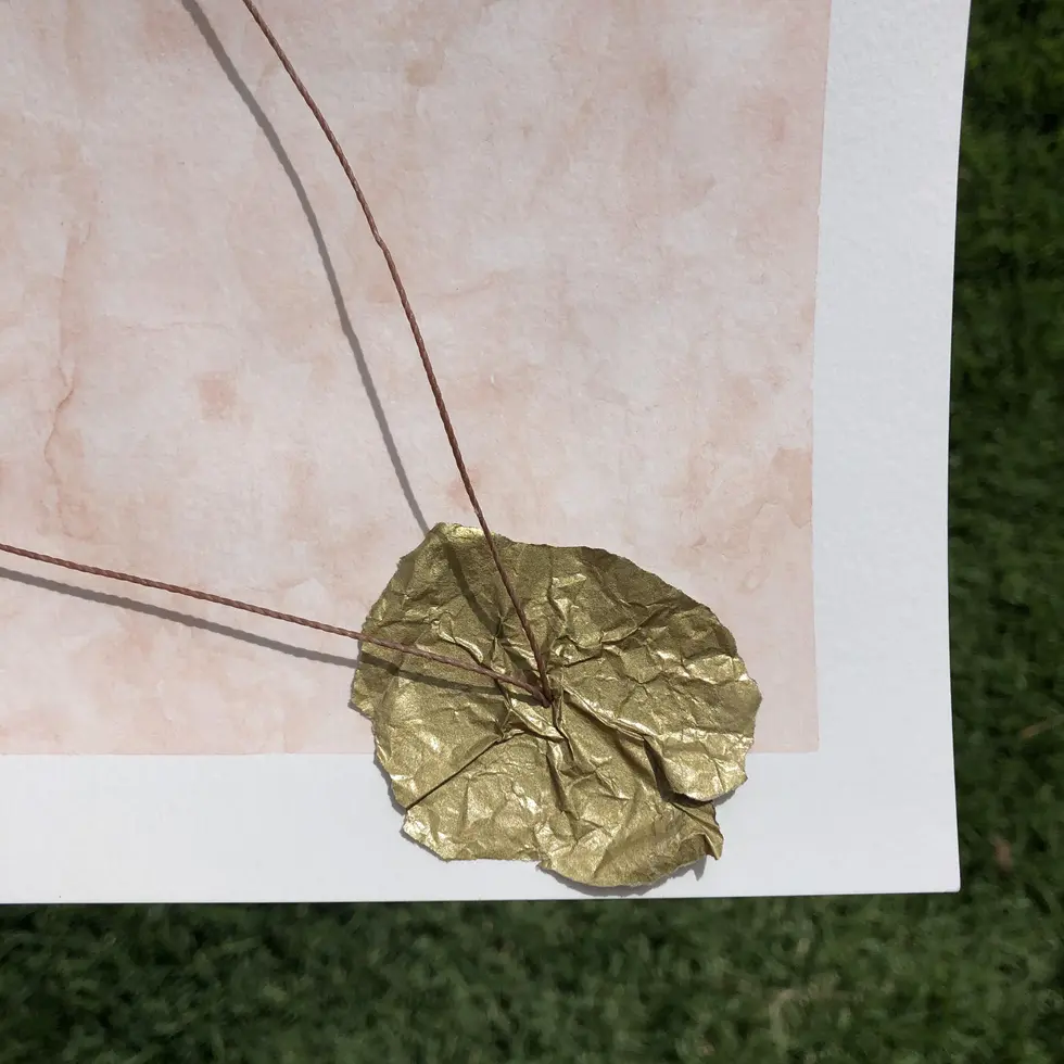 Colagem delicada com galho seco e fios dourados flutuando sobre fundo rosa névoa; mão segura composição com papéis texturizados, contraste com grama verde ao fundo.