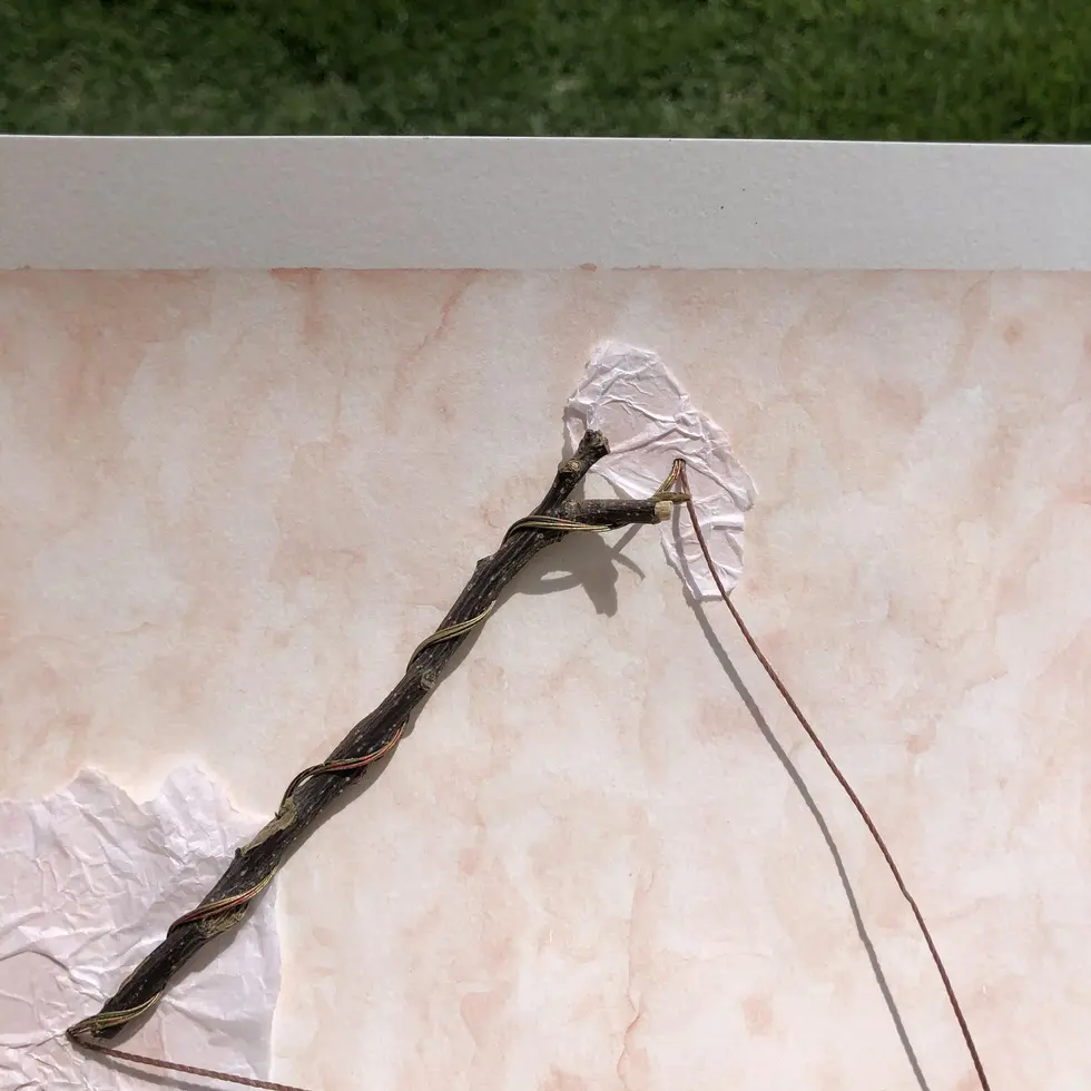 Colagem delicada com galho seco e fios dourados flutuando sobre fundo rosa névoa; mão segura composição com papéis texturizados, contraste com grama verde ao fundo.
