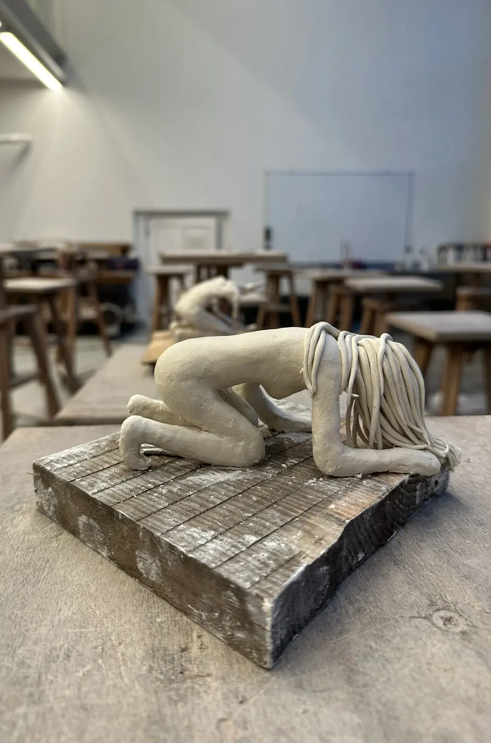 Escultura de figura humana em argila sobre base de madeira, corpo curvado com mãos e joelhos no chão, cabeça inclinada e cabelos caídos; postura de entrega e oração.
