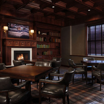 An interior photo of a reading room at the NorthPort Hotel. The room is cozy, with dark wood paneling, a fireplace with a roaring fire, and walls full of book shelves. 