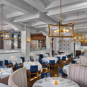 An interior photo of the dining room of the NorthPort Hotel. A room with approximately 10 dining tables and four booths. Tables and booths are set with place settings, have white table cloths, and there are brass reading lights in the centers. 