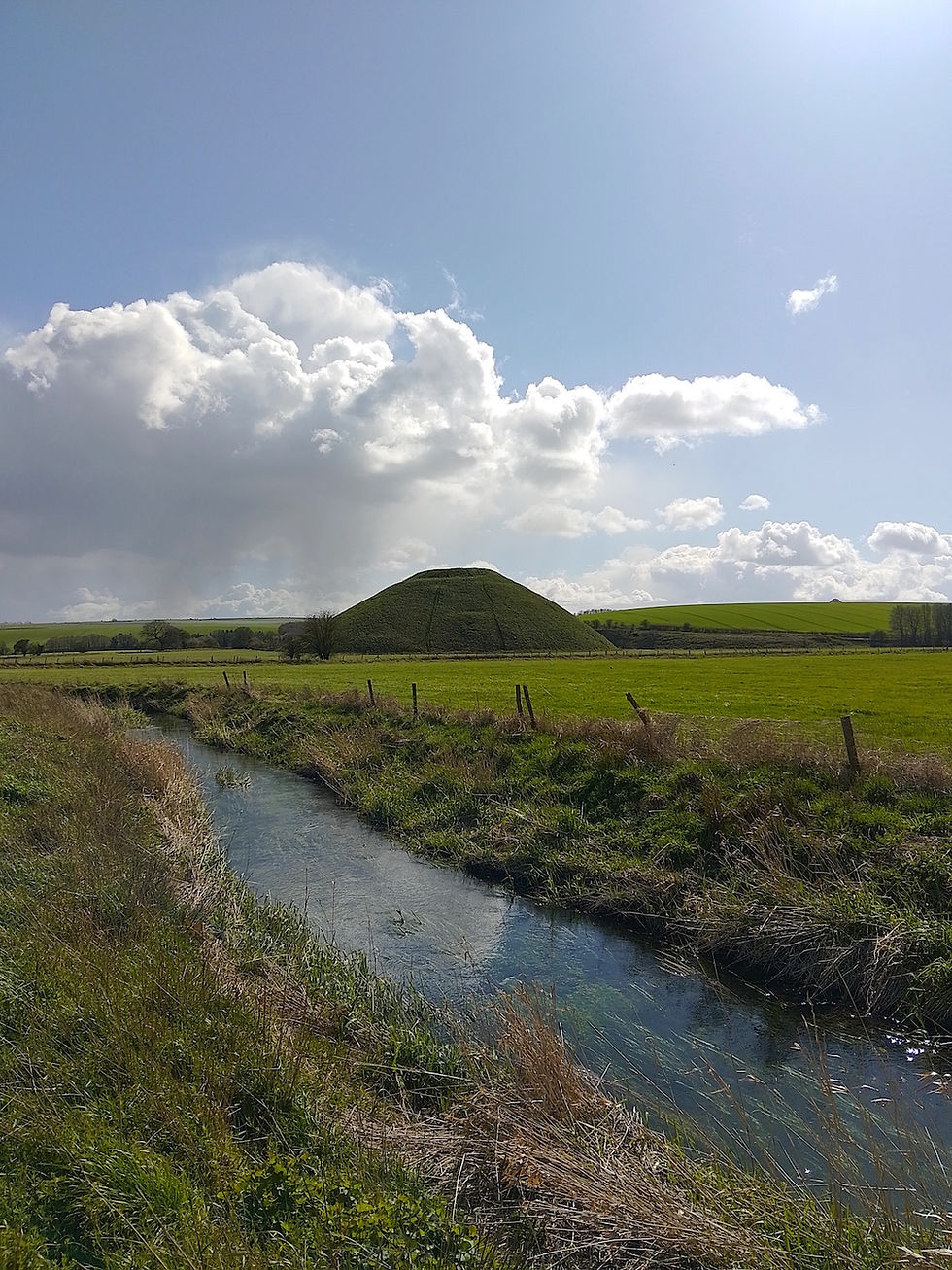 Silbury Hill under a blue sky with fluffy clouds. River Kennet flows through the green field, creating a calm and serene scene.