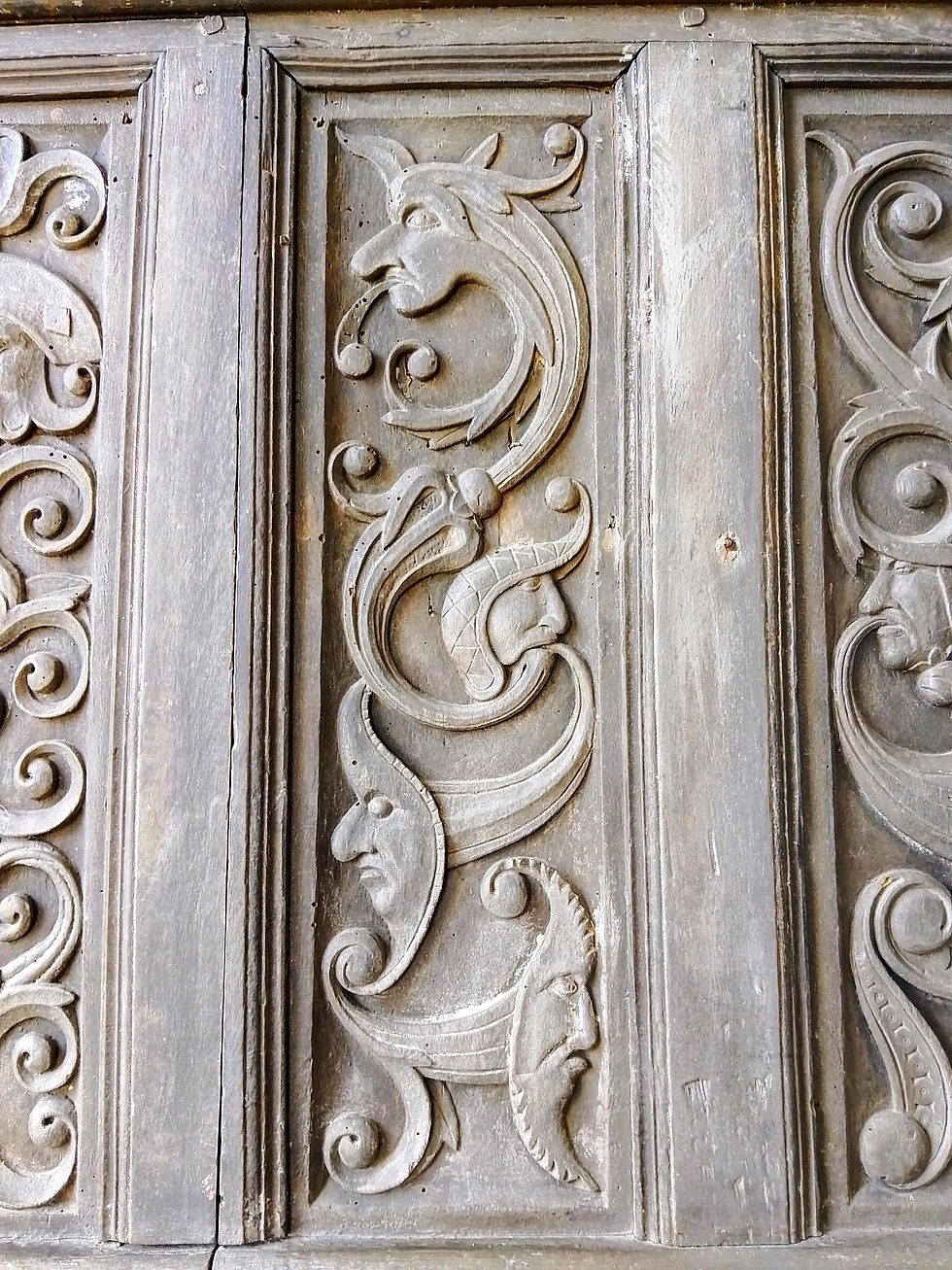 Intricately carved wooden panel featuring mythical faces and swirling patterns. The wood is aged with a rustic, detailed design.