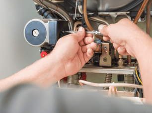 A person making a furnace repair with a screwdriver. The image focuses on hands working amidst pipes and metal parts. The setting is indoor.