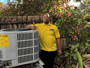 Lamar Werts, owner of The Service Pros, in yellow shirt gives thumbs up beside an air conditioning unit. HVAC service contractor. Wooden fence and green foliage in background. Sunny day, relaxed mood.