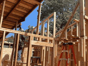 Construction workers building a wooden house frame under a clear blue sky. A red ladder is visible, with trees in the background. The image represents the construction of an ADU in St. Petersburg.