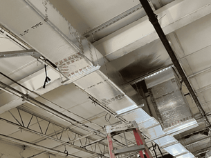 Ceiling with exposed metal beams and ventilation ducts, ladder visible below. Industrial setting, neutral colors, with wires hanging down. Representing duct installation for improved indoor quality in Tampa Bay.