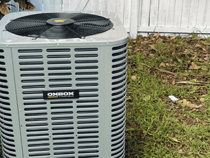 Air conditioning unit labeled "OXBOX" next to plants and a white fence; grass and leaves on the ground. This photo was taken by The Services Pros of a repair made with 10-year HVAC warranty.