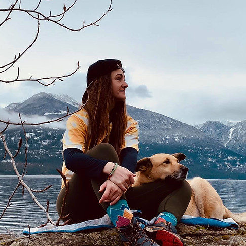 Jayce McKenzie sits on a huge rock with a yellow shirt and a backwards hat on the beach with moutains and Kootenay lake in the background. Her dog penny sits beside her with her head resting in her lap. They both look off into the distance.