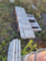 Stacked wooden planks, concrete slabs, and pallet on overgrown ground.