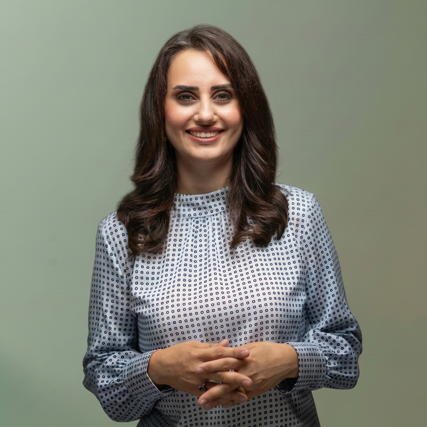 Smiling woman in a blue patterned blouse stands behind a pedestal against a muted green background.