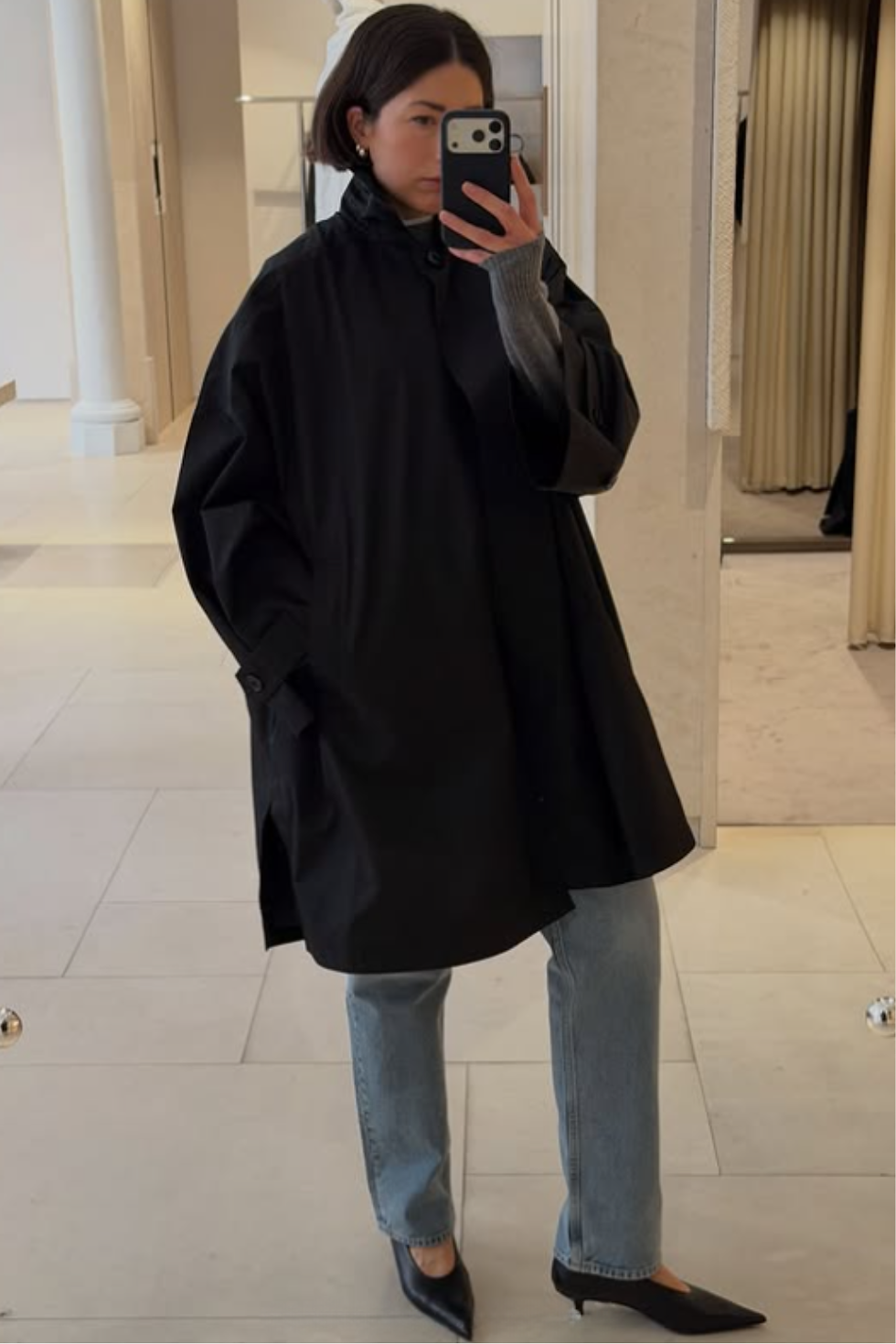 A woman taking a mirror selfie in a minimalist boutique, wearing black pointed-toe glove pumps with a low kitten heel. This 'Glove Pump' style is a featured Spring/Summer 2026 shoe trend representing Quiet Luxury. She has paired the heels with a black oversized coat, a grey sweater, and straight-leg light-wash jeans for an effortless Parisian look.