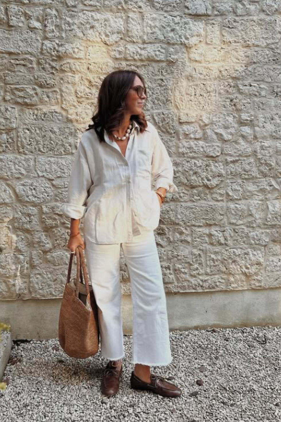 Woman in white outfit- white linnen shirt and white jeans- holds a brown woven bag, standing on gravel with a stone wall backdrop. She wears sunglasses and a necklace.