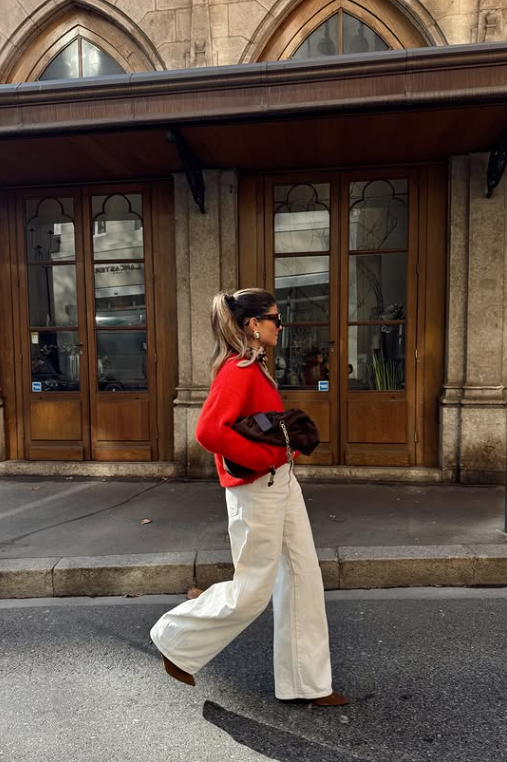 Woman in red sweater and white jeans walks on street holding brown purse. Background shows a building with arched windows and wooden doors.