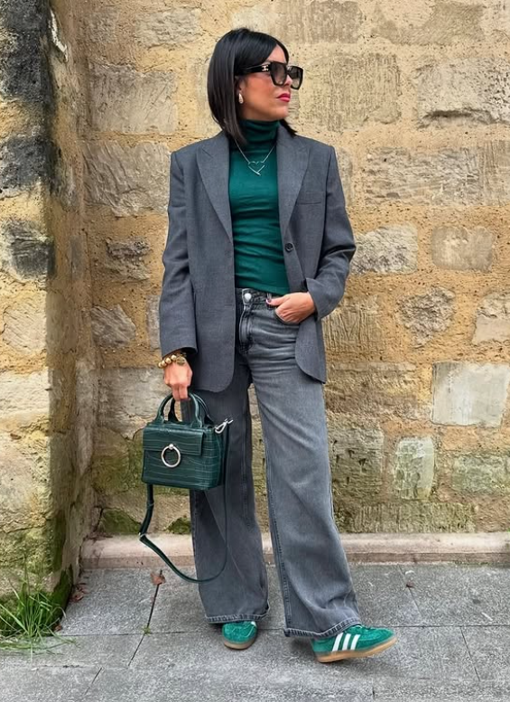 a woman wearing a green turtleneck, a green bag and sneakers, grey blazer and grey wide leg jeans and sunglasses.