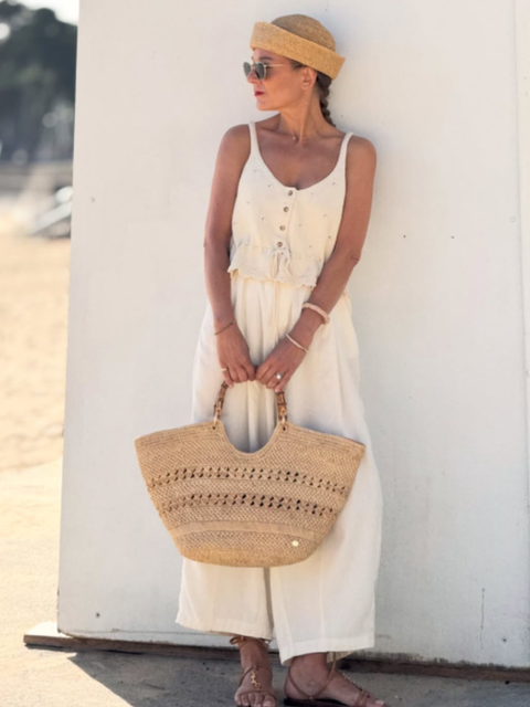 Woman in a cream sundress and straw hat stands by a white wall, holding a woven beach bag in warm sunlight.