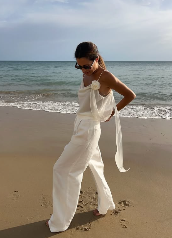 Chic summer holiday outfit with neutral tones – woman wearing a lightweight white top and wide-leg trousers on the beach, styled in a minimalist and effortless way.