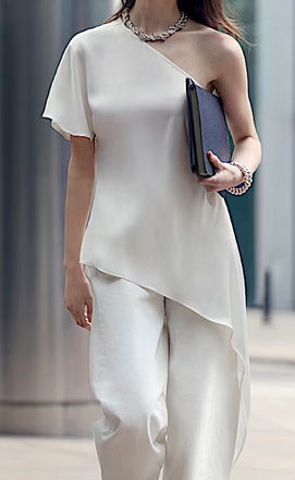 Woman in elegant white asymmetrical top and pants holds a black notebook. Urban setting with modern architecture in the background.