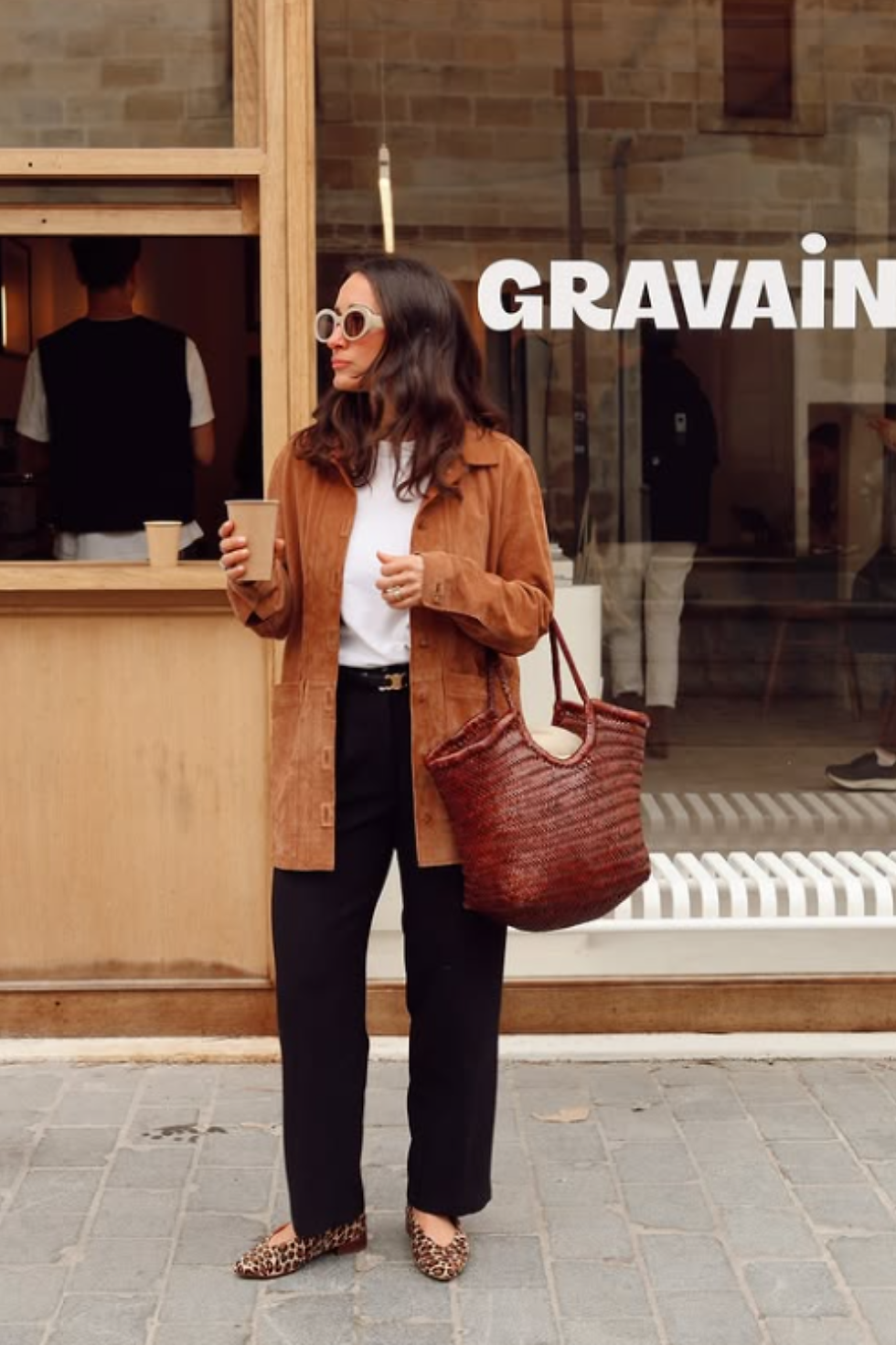 A woman standing outside a café wearing leopard print flat shoes, a key neutral shoe trend for Spring/Summer 2026. She is styled in a tan suede jacket, black trousers, and a large woven tote bag, demonstrating how to wear animal print shoes as a wearable everyday neutral.