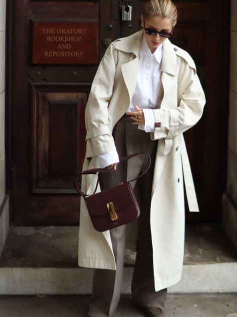 Woman in white trench coat, tailored trousers, classic loafers and sunglasses holding a burgundy bag, standing outside "The Oratory Bookshop and Repository" door.