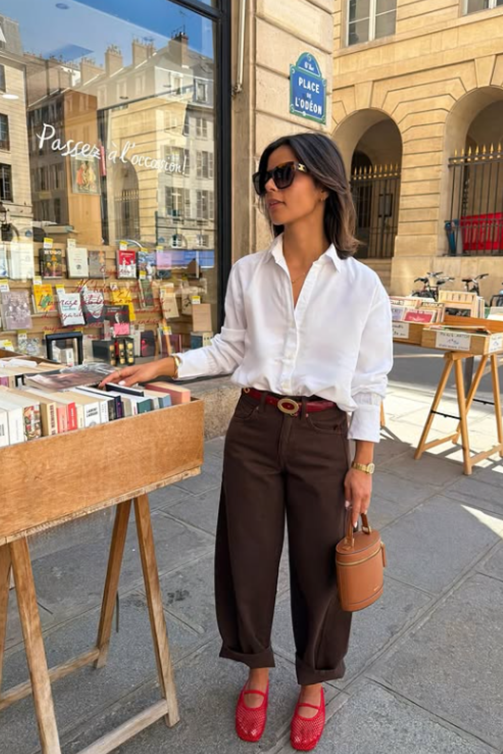 Woman in sunglasses browses books outside a Paris bookstall at Place de l’Odéon, wearing a white shirt, brown pants, and red shoes.