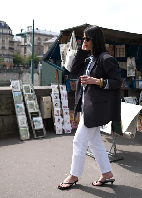 Woman in sunglasses walks by Parisian book stalls, holding coffee. She's in a black blazer, white jeans, black sandals. Calm city vibe.