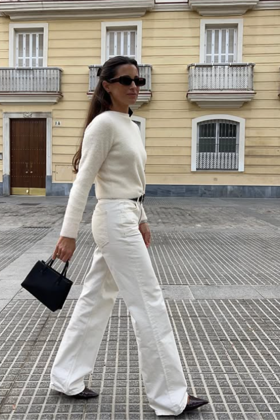Woman in creme knit and white jeans- a white monochrome outfit walks on patterned pavement, holding a black bag. Yellow building with balconies in background. Calm mood.