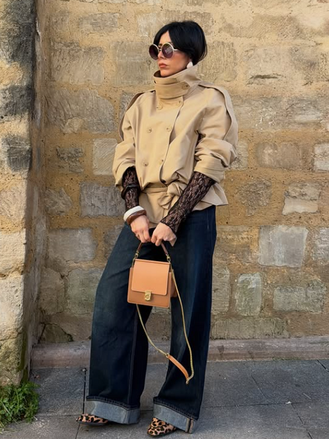 A person in a short beige trench coat and sunglasses stands against a stone wall holding a tan purse, exuding a chic, urban vibe.