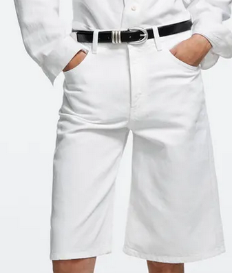 Person in white bermuda shorts and shirt with a black belt, hands in pockets, standing against a plain white background.