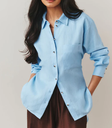 Woman in a light blue button-up shirt with hands in pockets, standing against a plain beige background.