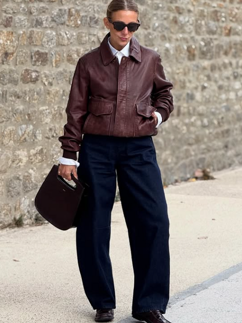 Chic minimalist outfit featuring a brown leather flight jacket, white button-down shirt, dark indigo wide-leg jeans, and burgundy loafers – how to style a leather jacket in 2026