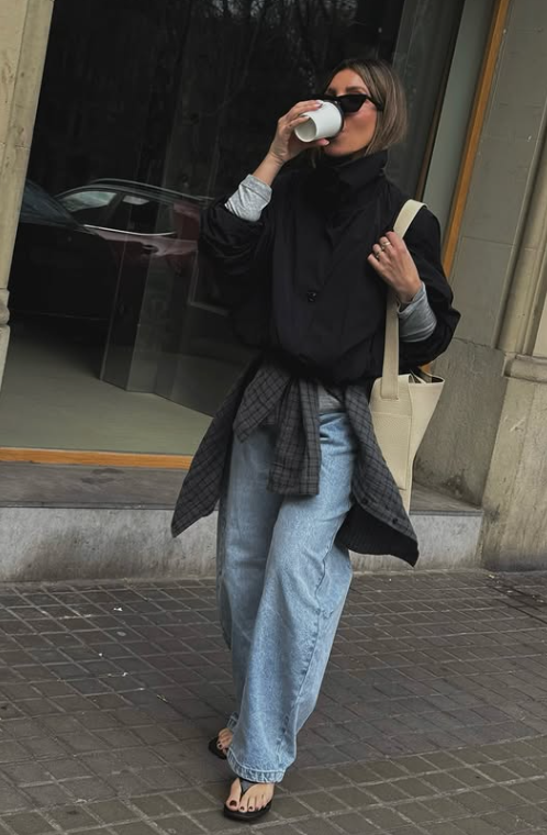 A woman standing on a city street in a Parisian-inspired casual outfit, wearing elegant black leather flip flops as part of the 'Fancy Flip Flops' Spring/Summer 2026 shoe trend. She is styled with wide-leg light-wash jeans, a black jacket with a plaid shirt tied at the waist, and a large cream tote bag, showcasing wearable elevated minimalism.