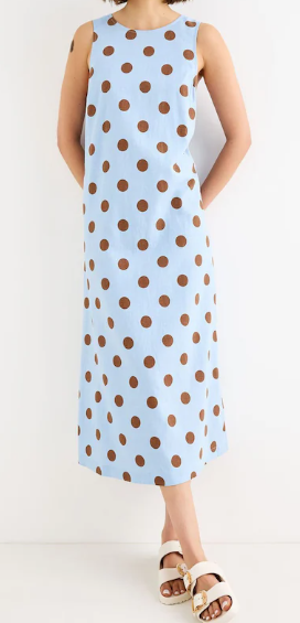 Woman modeling a light blue sleeveless maxi dress with brown polka dots, standing against a white background in white sandals.
