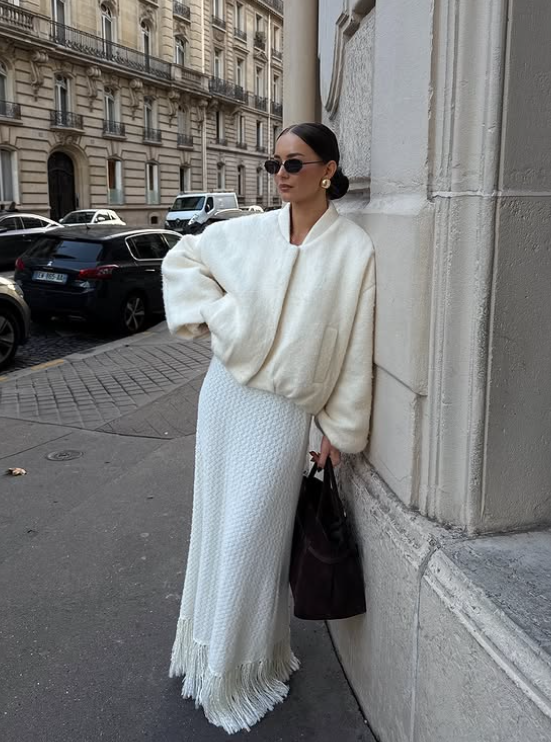 a woman wearing white ruffeled maxi skirt, creme bomber jacket, brown suede bag, statement gold earrings and sunglasses.