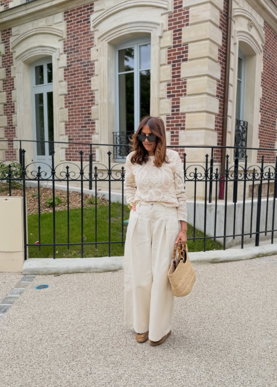 a woman wearing a creme crochet top with creme tailored pants and a raffia bag.