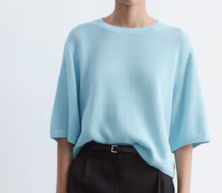 Person wearing a loose light blue top and black pants against a plain white background.